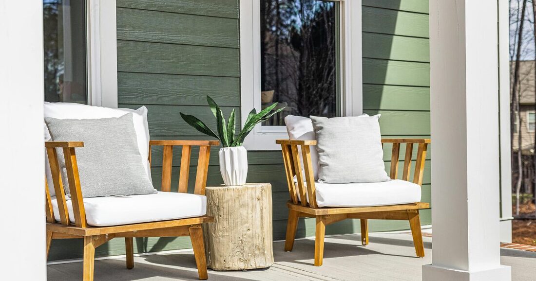 two wooden chairs with white cushions on a front porch of s green house it is sunny out