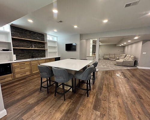basement remodel in south jersey with a bar and wooden cabinets. large island with bar chairs. flooring is brown LVP flooring in the bar area. in the couch and tv area there is a grey rug