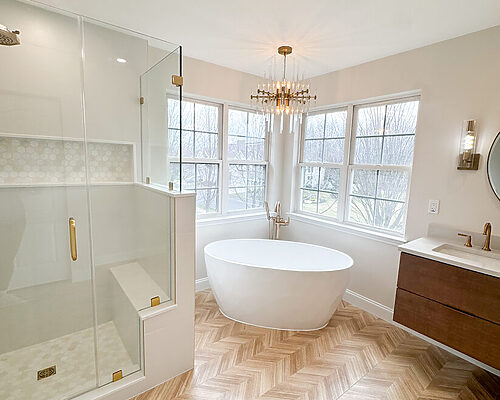 Modern bathroom remodel featuring a freestanding soaking tub beneath large windows, glass walk-in shower with brass fixtures, herringbone wood-look tile flooring, and a chandelier for a bright, spa-like feel.